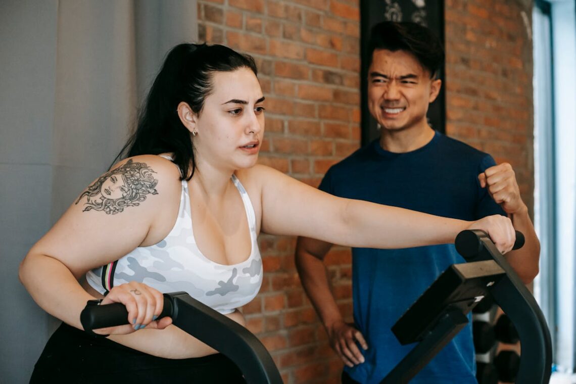 Exhausted young obese woman training on elliptical machine with support of Asian male instructor
